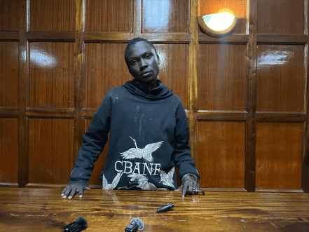 An image of a man standing behind a wooden table displaying firearms, captured for KWETU NEWS, highl.