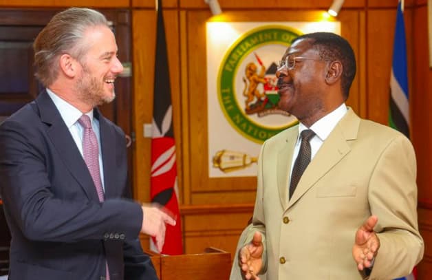 Kenyan and foreign officials exchanging greetings in a formal setting with national flags in the bac.