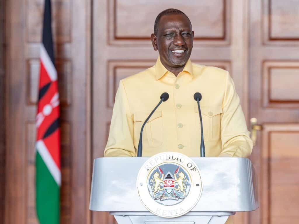 Kenyan official delivering a speech at a formal event with the national flag in the background.