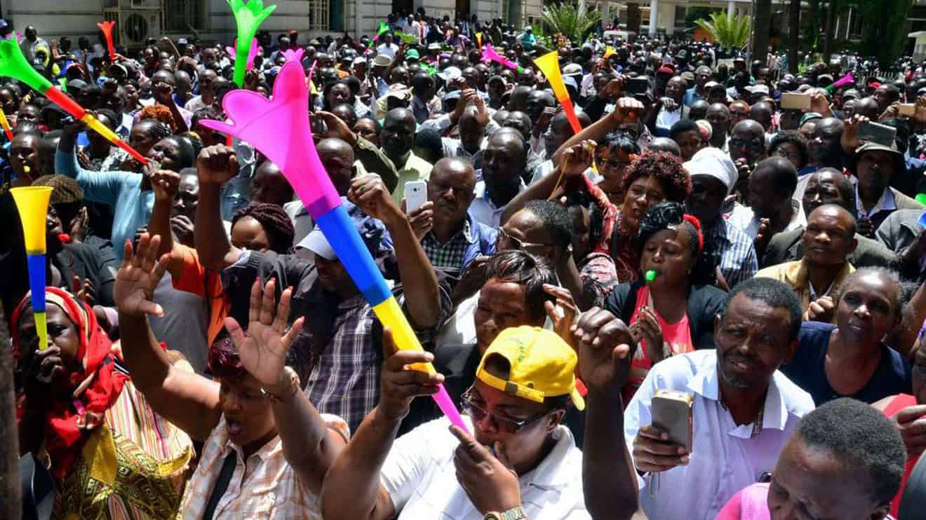 Large crowd of people celebrating outdoors with colorful balloons and waving hands.