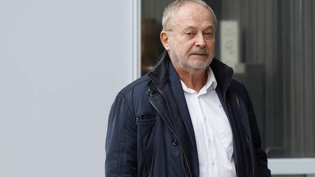 Senior man walking outdoors in urban environment, wearing a navy jacket and white shirt.