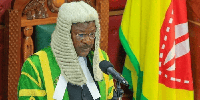 Senior judge delivering a speech at a courtroom, wearing traditional judicial robes and a white wig.