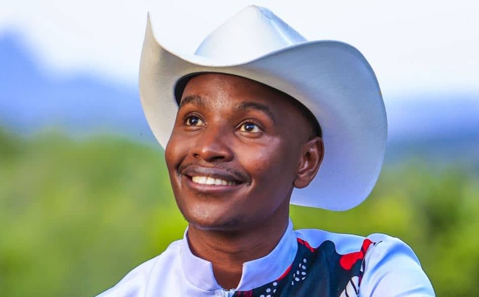 Kenyan man wearing a white cowboy hat and traditional attire smiling in a lush green landscape.