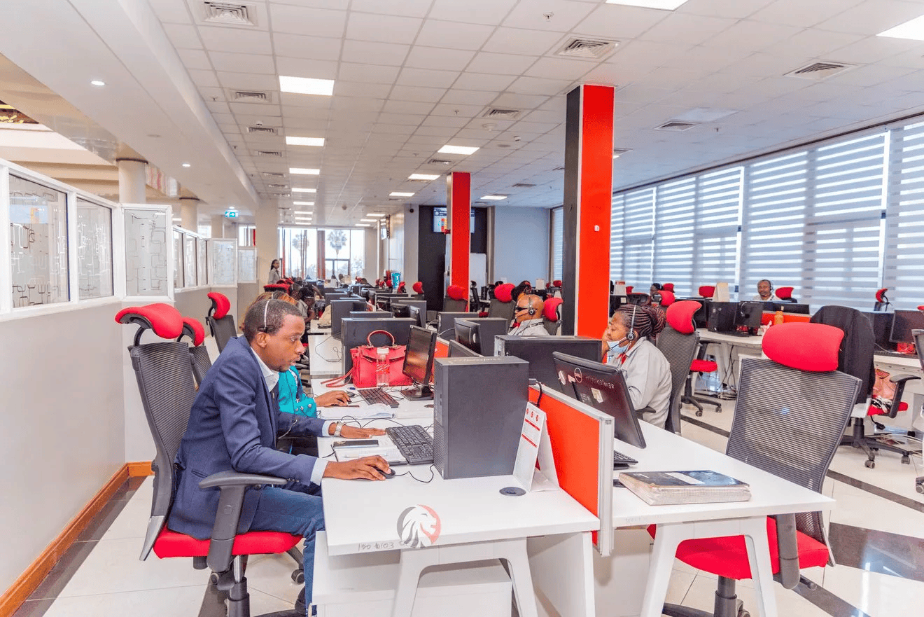 Employees working at desks in a modern, bright office with large windows and collaborative atmospher.